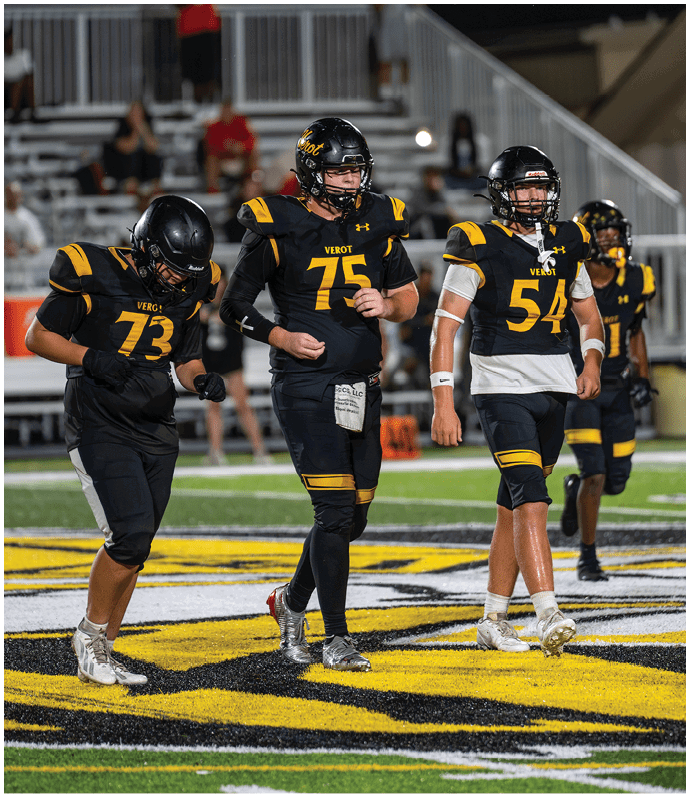 Two people wearing black and yellow football uniforms with helmets are on a field with blurred spectators in the background. Descripci n generada con IA