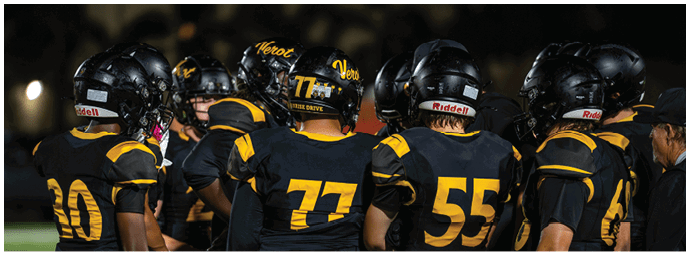 A group of people wearing black and yellow football uniforms and helmets are huddled together on a field at night. Descripci n generada con IA