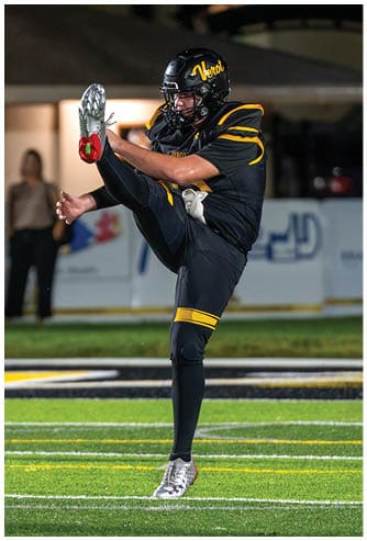 A person in a black and yellow football uniform is kicking a football on a field during a nighttime game. Descripci n generada con IA