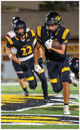 A person in a black and yellow football uniform is running with the ball while another person in a white and black uniform is falling behind on a football field. Descripci n generada con IA