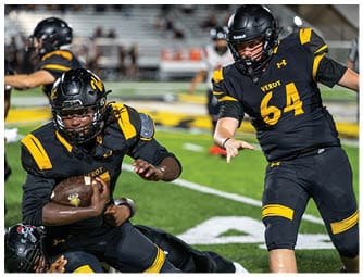 Two people in black and yellow football uniforms are on a field with one holding a football while the other reaches out, and a third person is on the ground behind them. Descripci n generada con IA