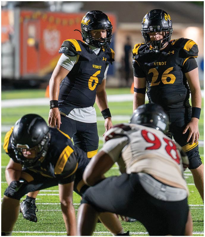 Two people in black and yellow football uniforms with helmets stand on a field facing two opponents in white and red uniforms during a game. Descripci n generada con IA
