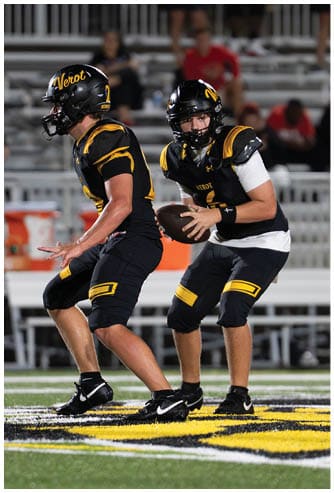 Two people in black and yellow football uniforms are on a field with one holding a football and a referee nearby while spectators sit in the background. Descripci n generada con IA