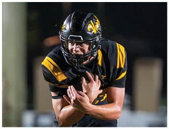 A person in a black and yellow football uniform is running with the ball on a field while two other players in different uniforms approach. Descripci n generada con IA