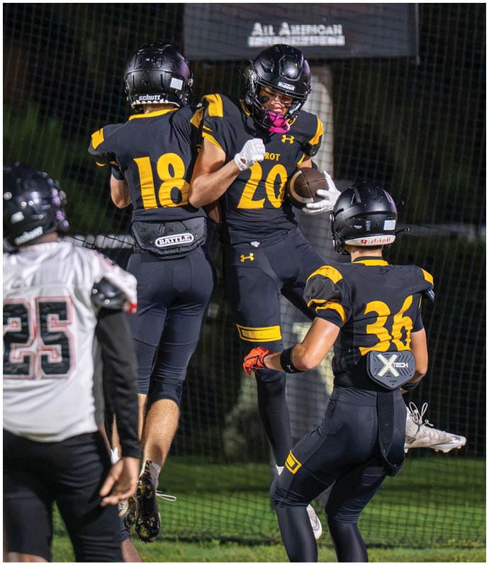 Three people in black and yellow football uniforms celebrate on a field at night with one holding a football. Descripci n generada con IA