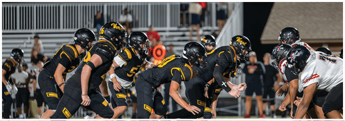 A group of people in black and yellow sports uniforms are lined up in a football formation facing a group in white and black uniforms on a field with a crowd in the background. Descripci n generada con IA