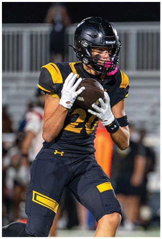 An adult wearing a black and yellow football uniform is catching a football on a field during a game. Descripci n generada con IA