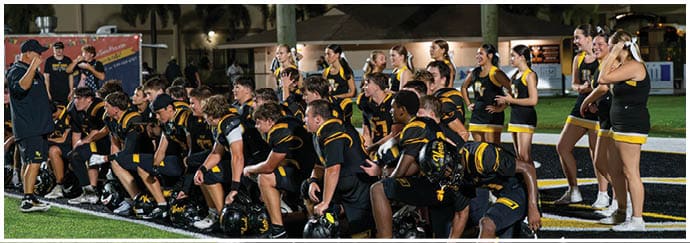 A group of people wearing black and yellow sports uniforms are gathered on a field, some kneeling and others standing, appearing to listen to a person speaking to them. Descripci n generada con IA