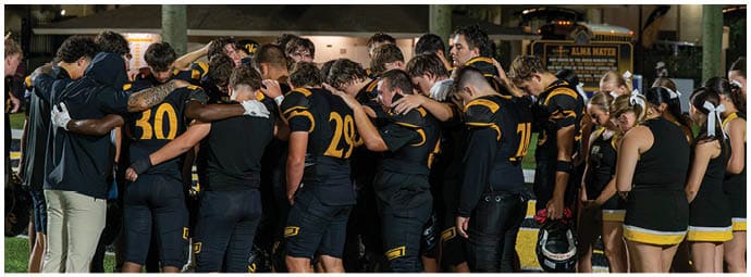 A group of people wearing black and yellow sports uniforms stand closely together in a huddle outdoors at night. Descripci n generada con IA