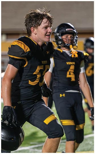 A person with wet, tousled hair wearing a black and yellow football uniform is smiling and looking to the side on a sports field. Descripci n generada con IA