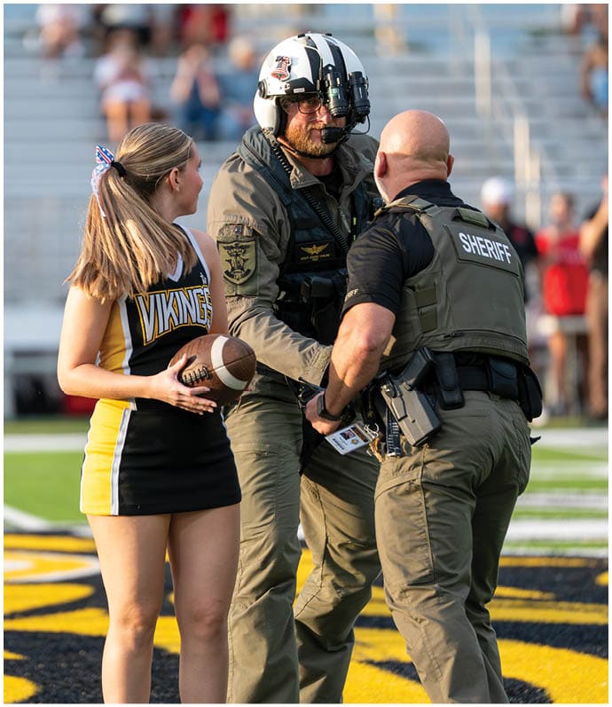A person in a Vikings cheerleader outfit holds a football while standing next to two people in tactical gear, one wearing a helmet and the other a vest labeled sheriff, on a sports field with blurred spectators in the background. Descripci n generada con IA