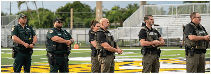 Five adults wearing tactical or law enforcement uniforms stand in a line on a sports field with their hands clasped in front of them. Descripci n generada con IA