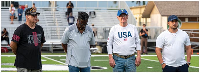 Four adults stand on a sports field with bleachers and other people in the background, wearing casual clothing including a USA sweatshirt and caps. Descripci n generada con IA