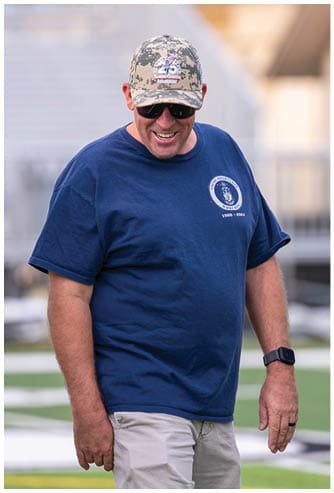 A person wearing a camouflage cap, dark sunglasses, and a navy blue t-shirt with a circular emblem is smiling outdoors. Descripci n generada con IA