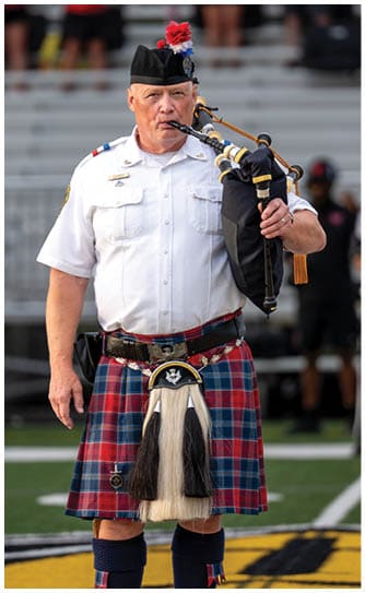 A person wearing a traditional Scottish kilt and uniform is playing bagpipes on a sports field. Descripci n generada con IA