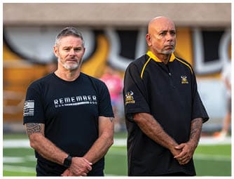 Two adults stand side by side on a sports field wearing casual athletic clothing with serious expressions. Descripci n generada con IA
