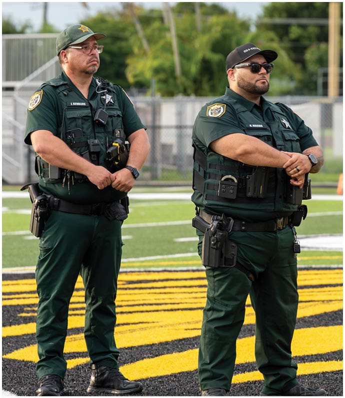 Two adults wearing green uniforms and tactical gear stand on a sports field with a yellow and black design on the ground. Descripci n generada con IA