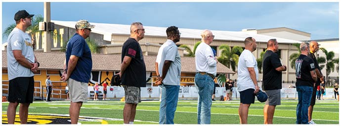 A group of adults standing in a line on a sports field, facing to the right, with some wearing casual clothing and hats. Descripci n generada con IA
