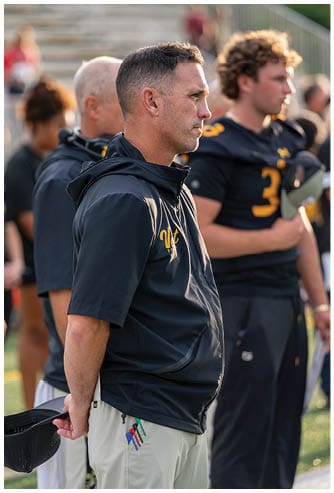 A person wearing a dark hooded jacket with yellow lettering stands in profile with two other people in dark sports uniforms blurred in the background. Descripci n generada con IA
