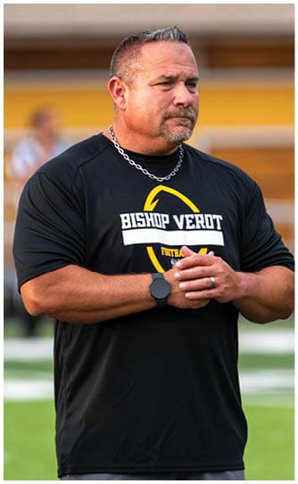 A person wearing a black Bishop Verot football shirt and a silver chain necklace stands on a sports field with hands clasped, looking focused. Descripci n generada con IA