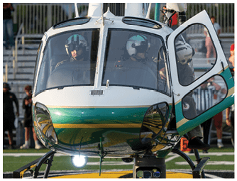 A helicopter is landed on a sports field with several people standing in the background near bleachers. Descripci n generada con IA