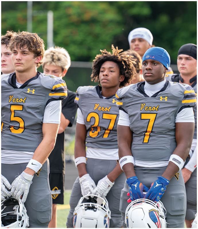 A group of people wearing gray and yellow football uniforms stand in a line on a field holding their helmets. Descripci n generada con IA