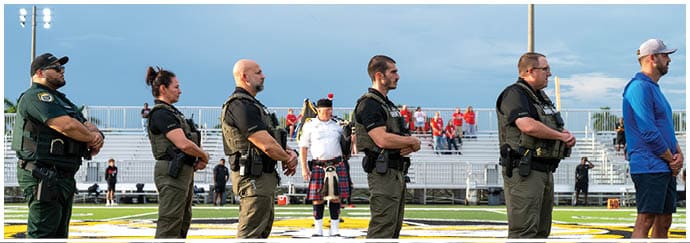 Seven people stand in a line on a sports field, six wearing uniforms with tactical gear and one in a blue shirt and shorts, with a person in a ceremonial kilt outfit standing behind them. Descripci n generada con IA
