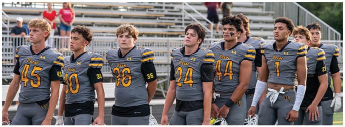 A group of adults wearing gray and yellow football uniforms stand in a line on a field holding white helmets. Descripci n generada con IA