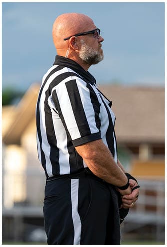 A person wearing a black and white striped referee uniform stands with hands clasped in front on a sports field. Descripci n generada con IA