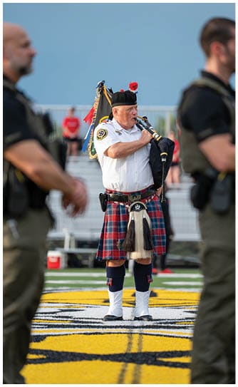 A senior person wearing a traditional Scottish outfit plays the bagpipes outdoors with blurred spectators in the background. Descripci n generada con IA