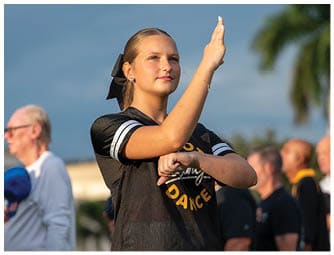 A person with a ponytail and a black bow is performing a dance move outdoors while several other people stand in the background. Descripci n generada con IA