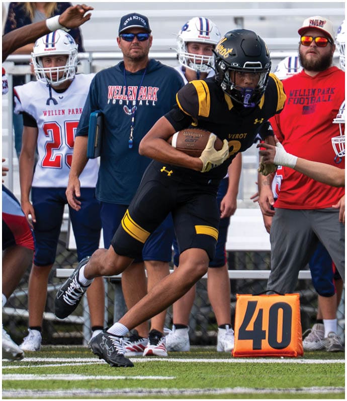 A person in black and yellow football gear is running with the ball while another person in white football gear reaches out to tackle them on a field with coaches and players in the background. Descripci n generada con IA