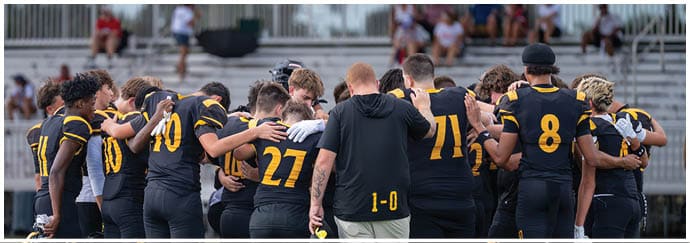 A group of people wearing black and yellow sports uniforms are huddled together in a circle on a field with blurred spectators in the background. Descripci n generada con IA