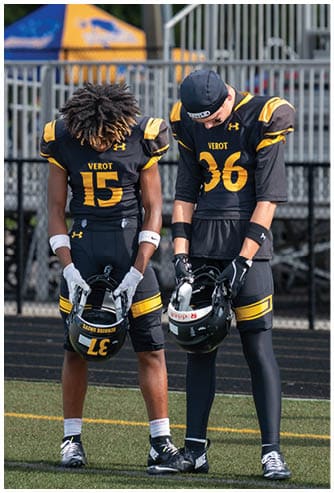 Two people in black and yellow football uniforms stand on a field holding their helmets and looking down. Descripci n generada con IA