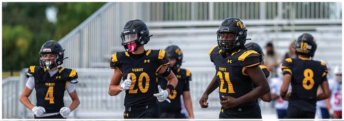 Four people in black and yellow football uniforms and helmets are standing on a field with bleachers in the background. Descripci n generada con IA