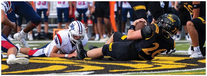 A group of people in football uniforms are engaged in a play on a field with one person lying on the ground holding the ball while others surround them. Descripci n generada con IA