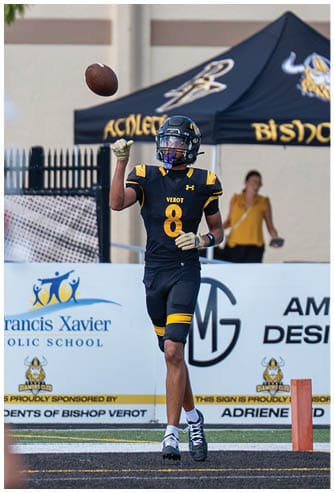 A person in a black and yellow football uniform with the number 8 is catching a football near a goal post with a tent and banners in the background. Descripci n generada con IA