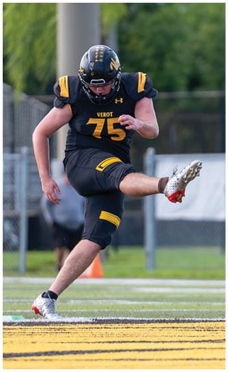 A person wearing a black and yellow football uniform is kicking a football on a field with a blurred background of trees and fencing. Descripci n generada con IA