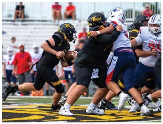 A group of people in football uniforms are actively engaged in a play on a football field with one person holding the ball and others blocking or tackling. Descripci n generada con IA