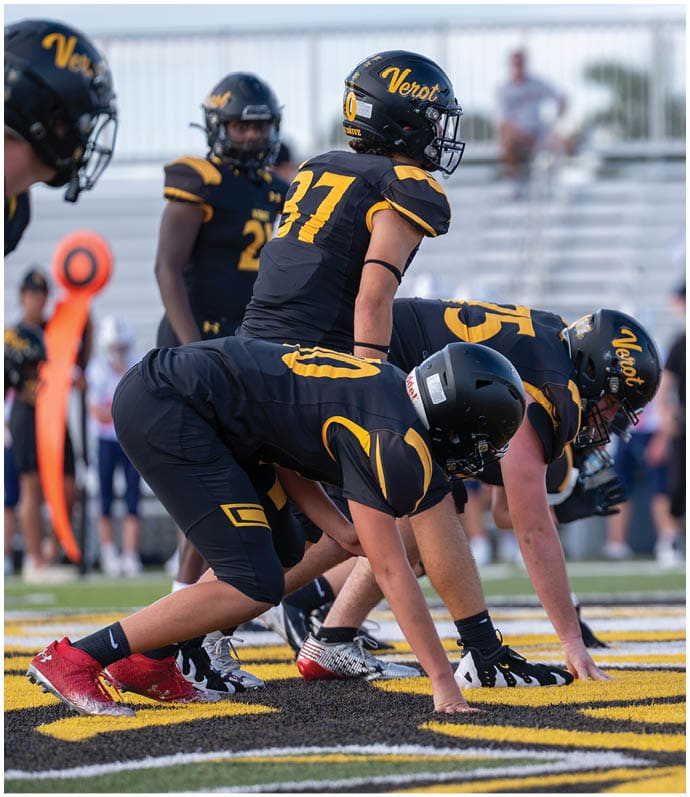 Several people in black and yellow football uniforms are positioned on a field facing an opponent holding a football, preparing for a play. Descripci n generada con IA
