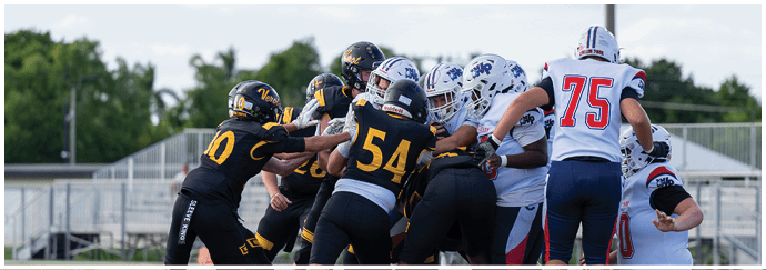 People wearing football uniforms are engaged in a close, physical play on a field with a blurred background of trees and bleachers. Descripci n generada con IA