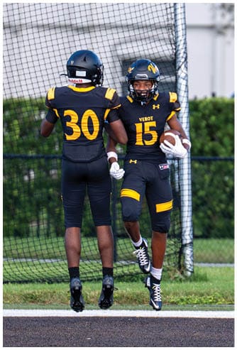 Two people in black and yellow football uniforms are holding hands, with one holding a football and both standing on grass in front of a net. Descripci n generada con IA