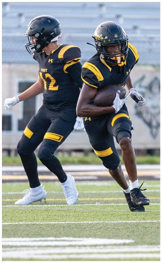 Two people in black and yellow football uniforms are on a field, with one person holding a football and the other preparing to move. Descripci n generada con IA