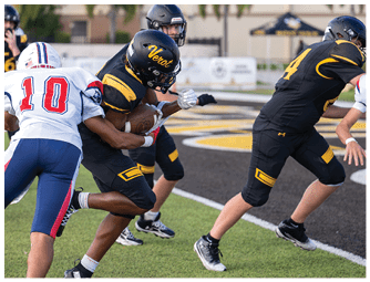 People in football uniforms are actively engaged in a play on a field with one person holding the ball while others attempt to tackle or block. Descripci n generada con IA