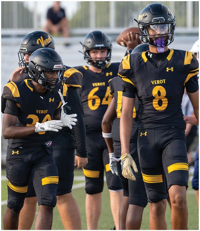 Several people wearing black and yellow football uniforms with helmets are standing on a field, appearing to be engaged in conversation or preparing for a game. Descripci n generada con IA