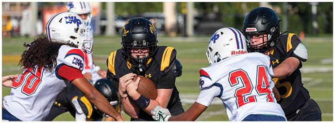 People wearing football uniforms are actively engaged in a play on a grassy field with one person holding the ball while others attempt to block or tackle. Descripci n generada con IA