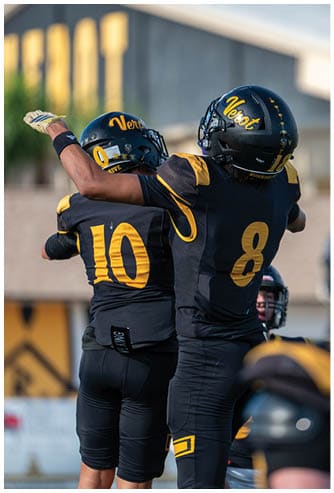 A person wearing a black and yellow football helmet and uniform raises one arm while standing outdoors near a blurred building. Descripci n generada con IA