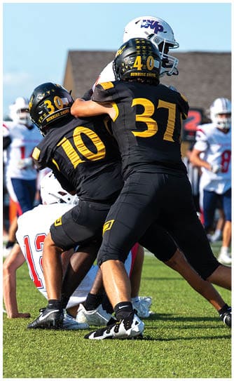 Two people in black and yellow football uniforms are engaged in a play on a green field while several people in white and blue uniforms stand in the background. Descripci n generada con IA