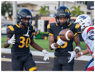 A person wearing a black and yellow football uniform and helmet is holding a football while another person in a white helmet and uniform stands nearby. Descripci n generada con IA