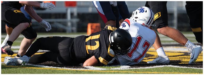 Two people in football uniforms are on the ground during a game while other players stand nearby on the field. Descripci n generada con IA